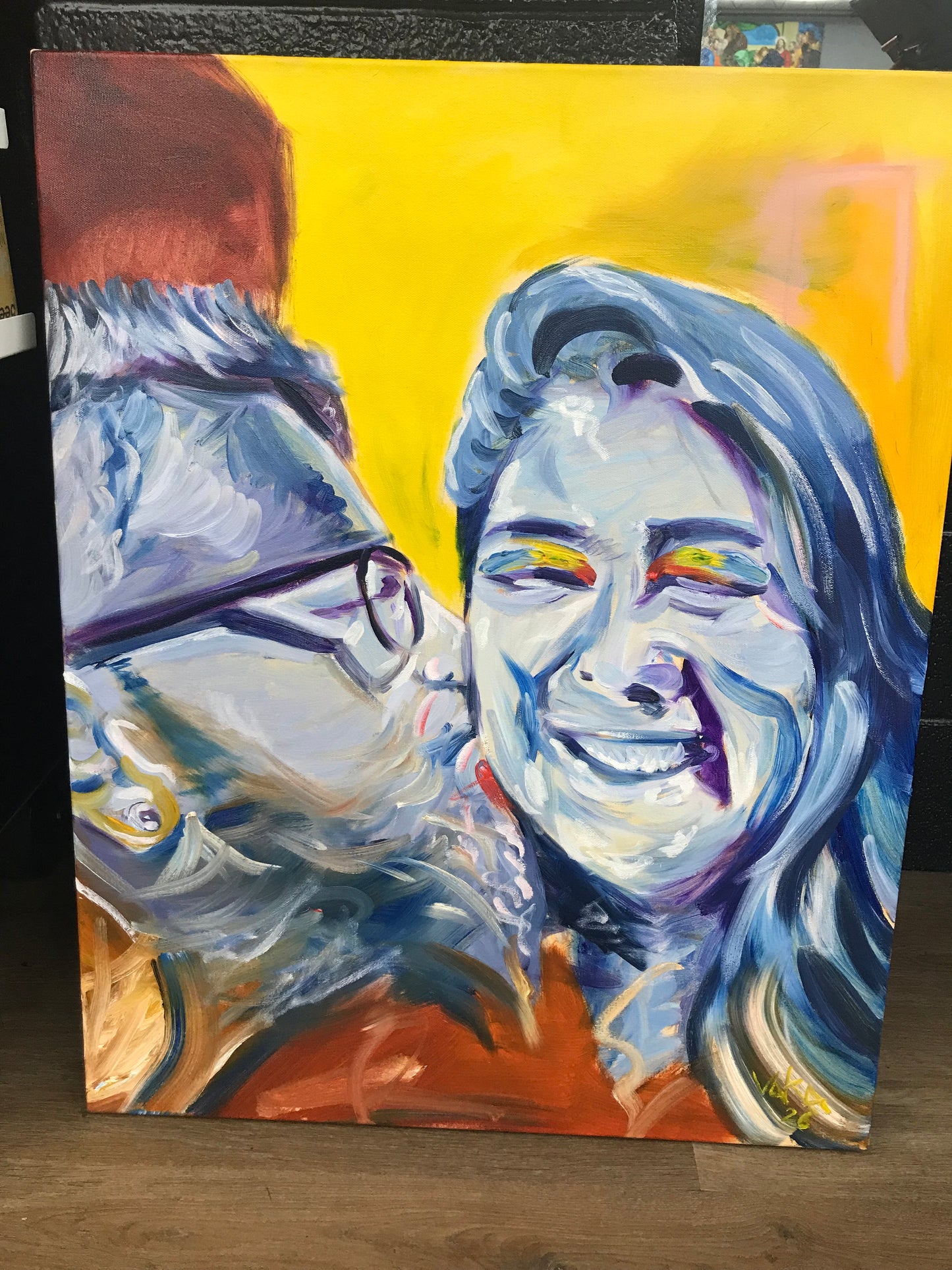 An expressive oil painting of two embracing figures, featuring bold brushstrokes in blue, yellow, orange, and white, displayed on a wooden surface.