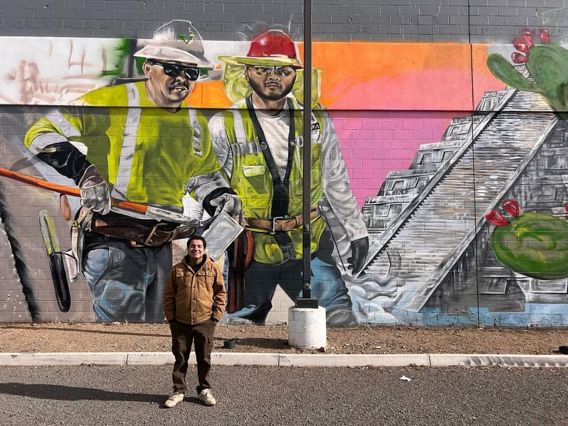 Person standing in front of a mural of construction workers and a colorful abstract design.