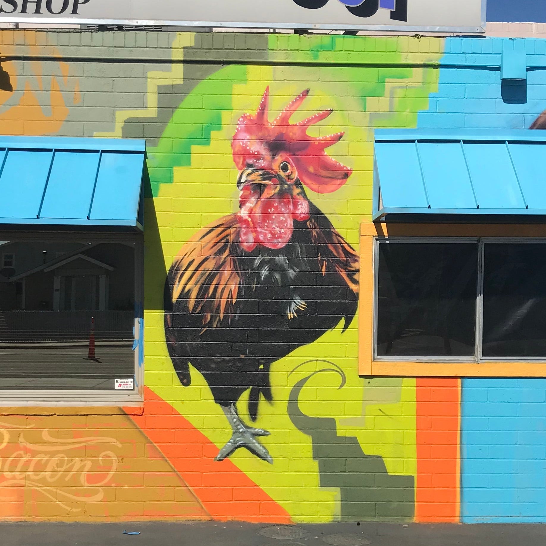 Colorful mural of a rooster on a building wall with a visible brand name above.