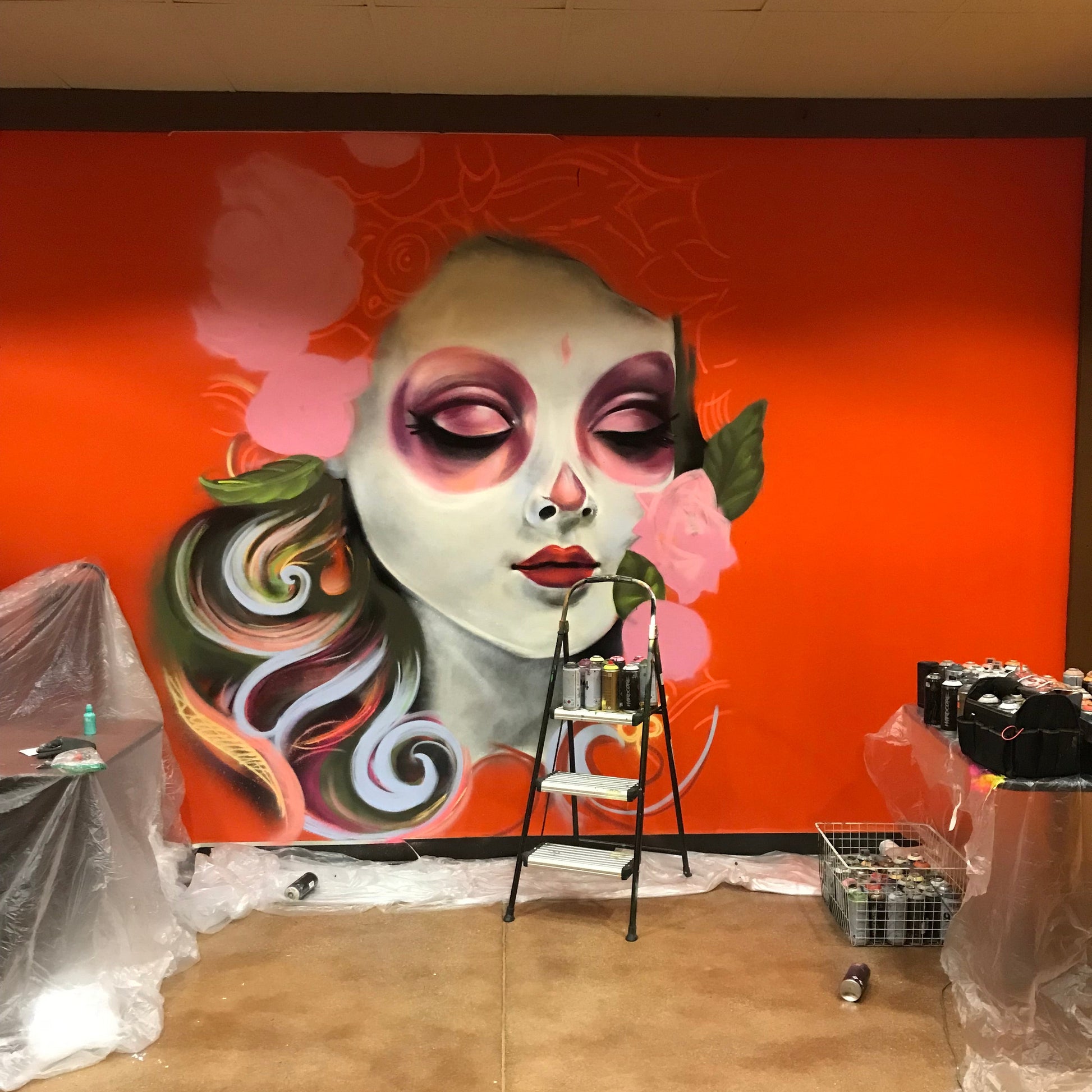 Mural of a stylized face with colorful hair on an orange background, with art supplies and a ladder in the foreground.